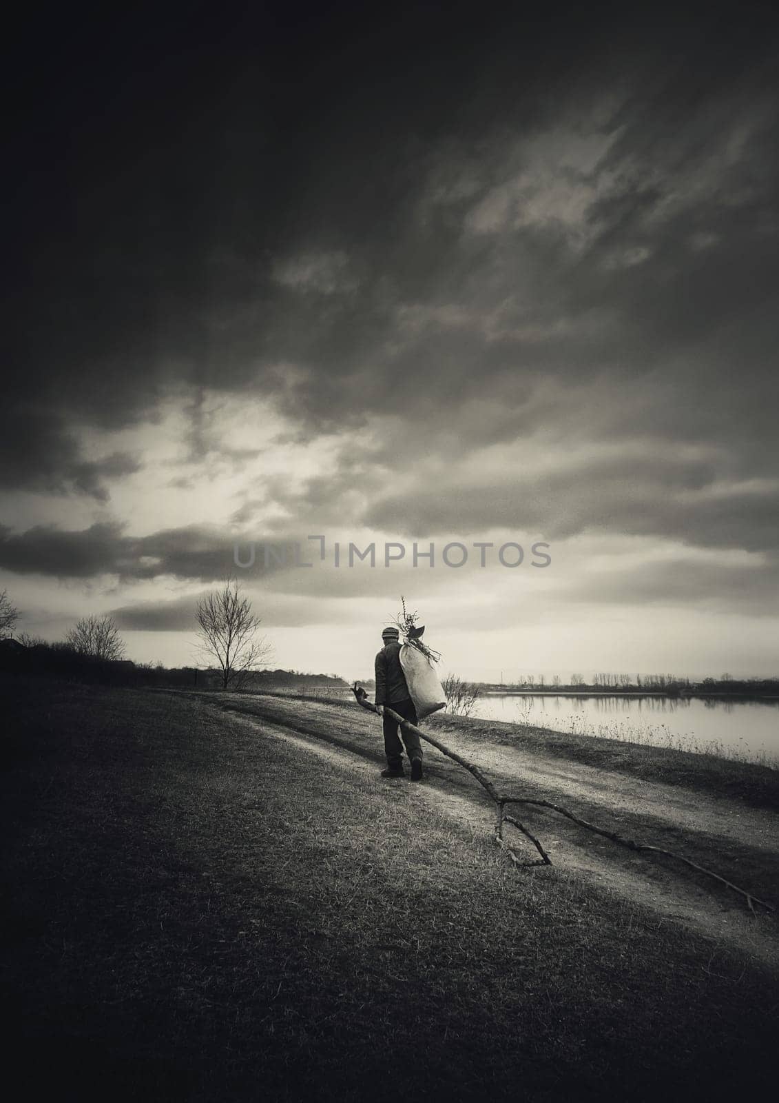 Lonely nomad man carrying firewood for heating, walking a country road. Moody and dramatic scene, black and white vertical background