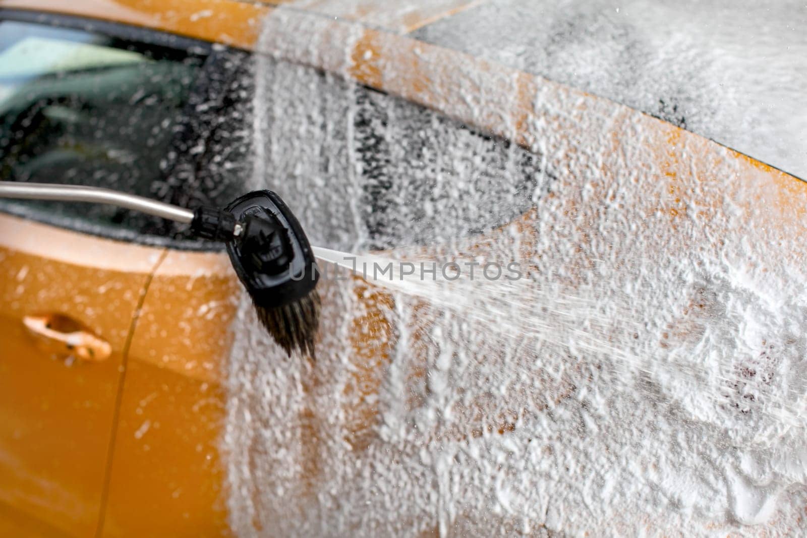 Shampoo and foam being sprayed on yellow car side, cleaned at carwash.