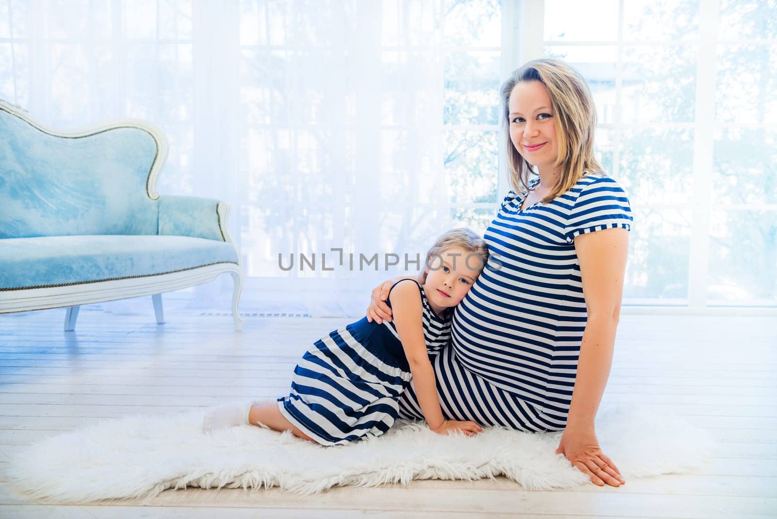 Beautiful pregnant woman with daughter sitting near window at home