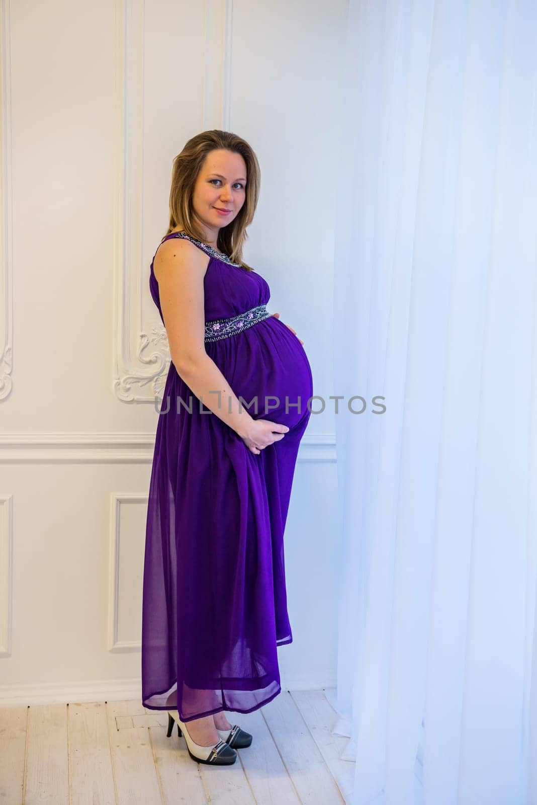 Beautiful pregnant woman in an ultraviolet dress standing near the window.