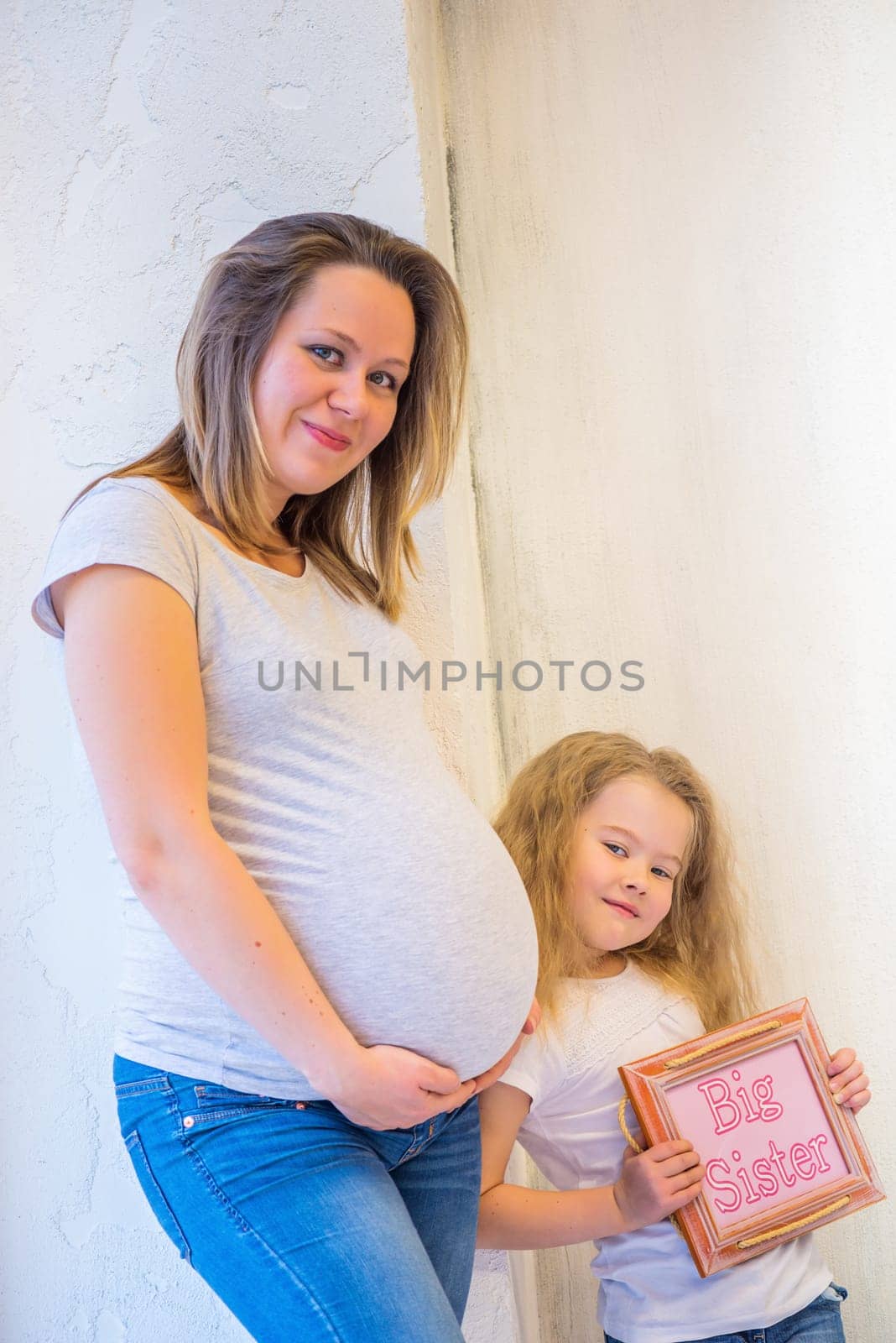 Beautiful pregnant woman with daughter standing near wall at home.