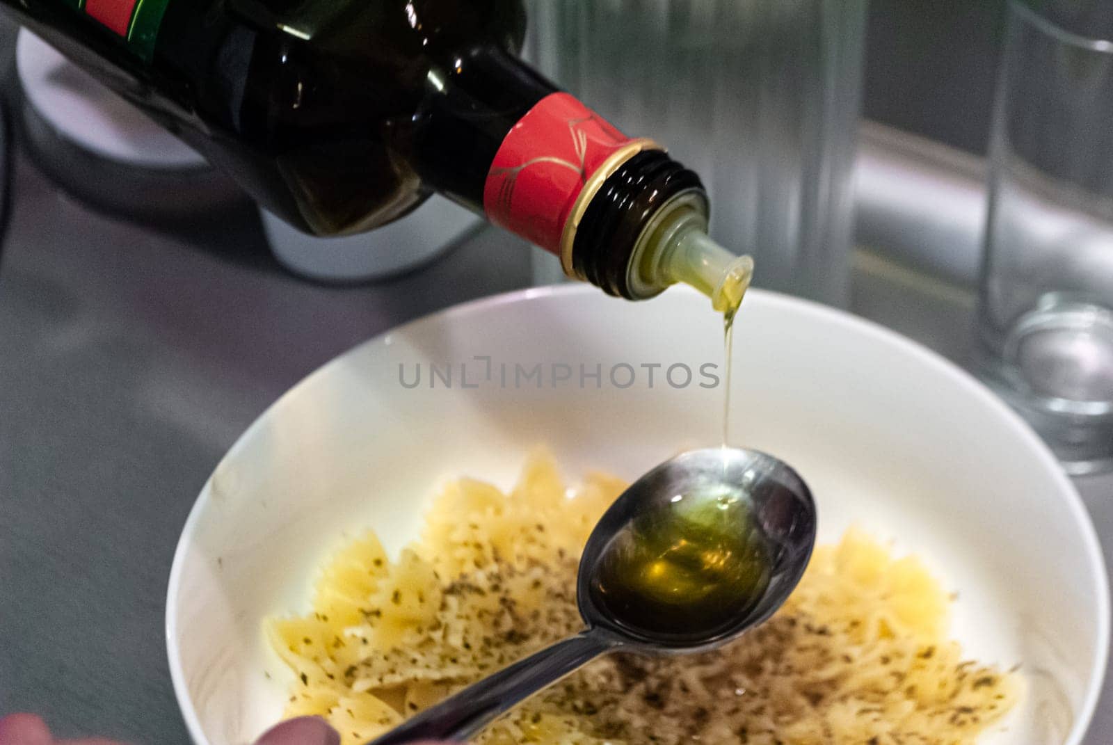 Step by step home cooking salad recepie. Olive oil pours into a spoon against the background of a bowl with pasta