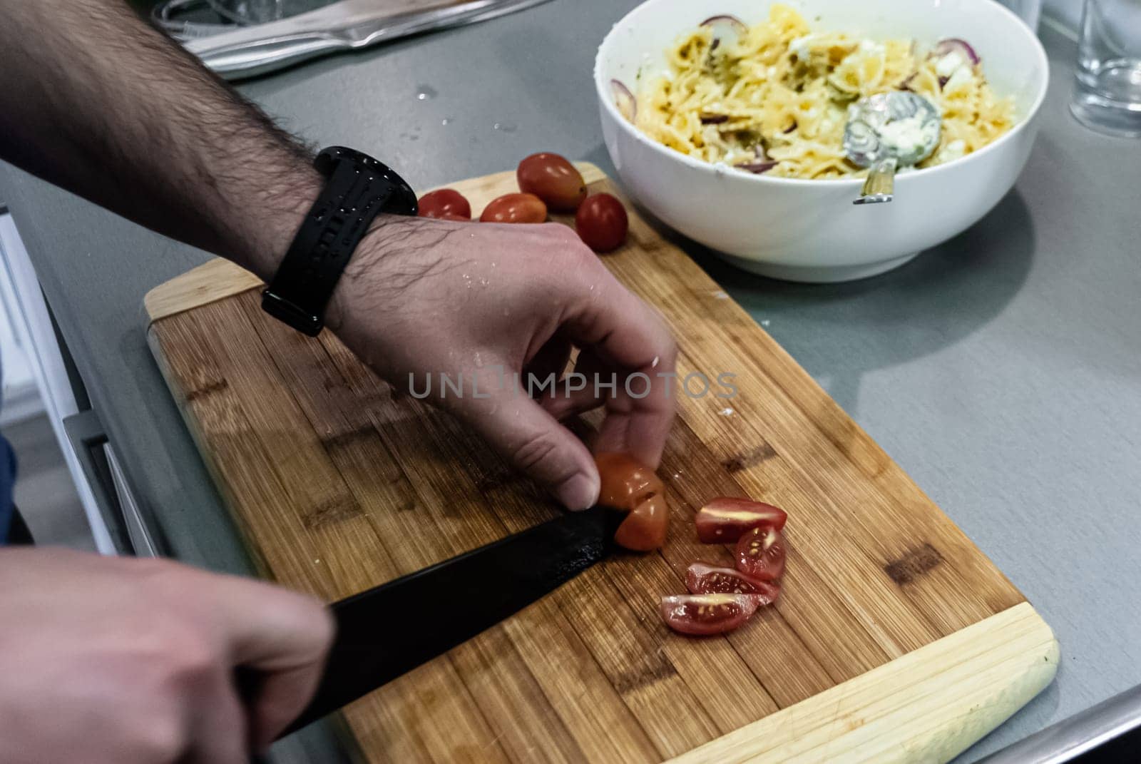 Step by step home cooking salad recepie. A man cuts cherry tomatoes on a wooden board against a white bowl of pasta