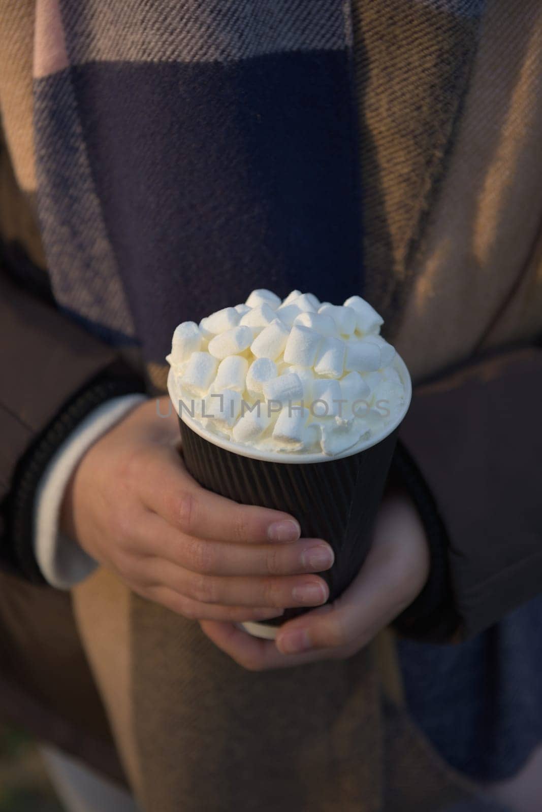 Vertical image of young and youthful female hands holding a hot chocolate to go in a black and white paper cup. Earthy color winter clothes background