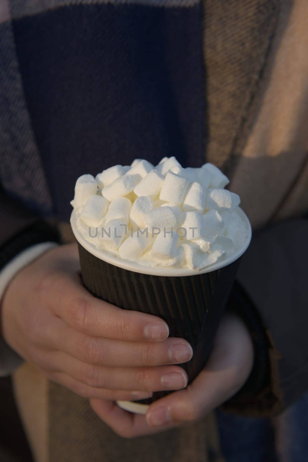 Hot take away beverage with an abundance of marshmallows and cream on top, cradled by youthful female hands. Likely hot chocolate, coffee or tea.