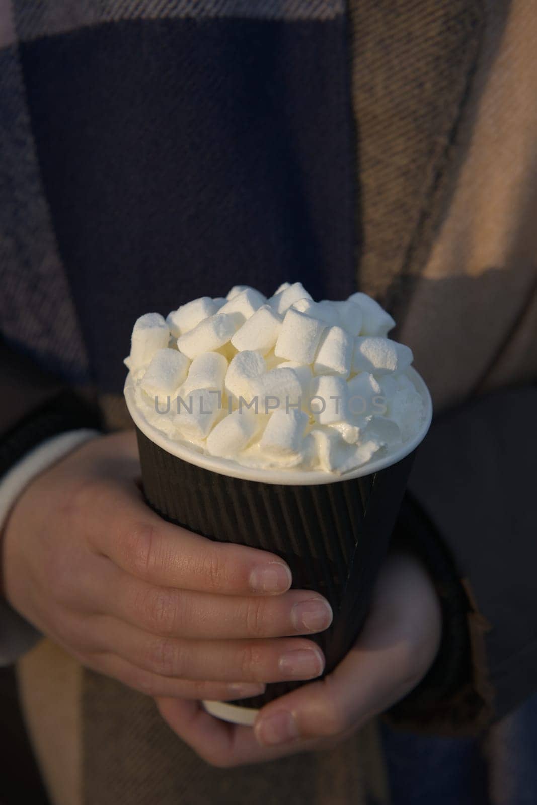 Close up vertical image of a young woman warming her hands on a freshly prepared takeaway hot drink. Winter scarf background, copy space above