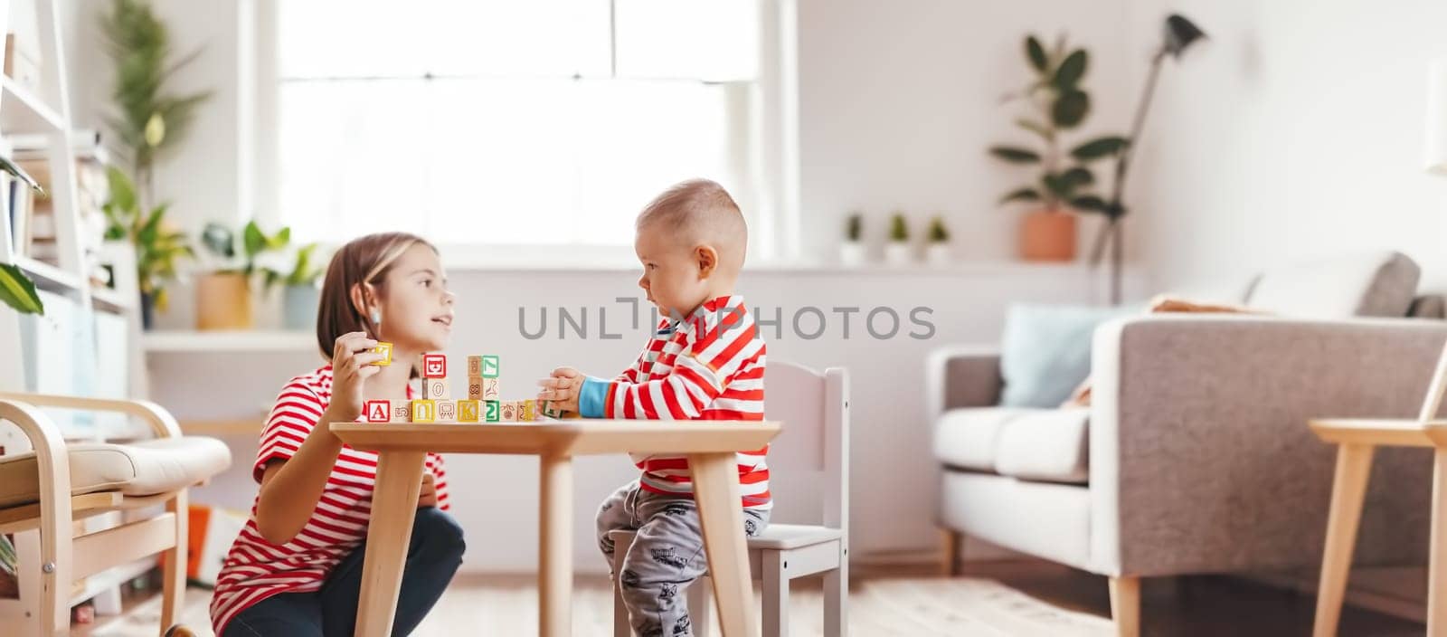 Sister teaching little brother letters by using cubes with symbols indoors. Concept of the family relationship.