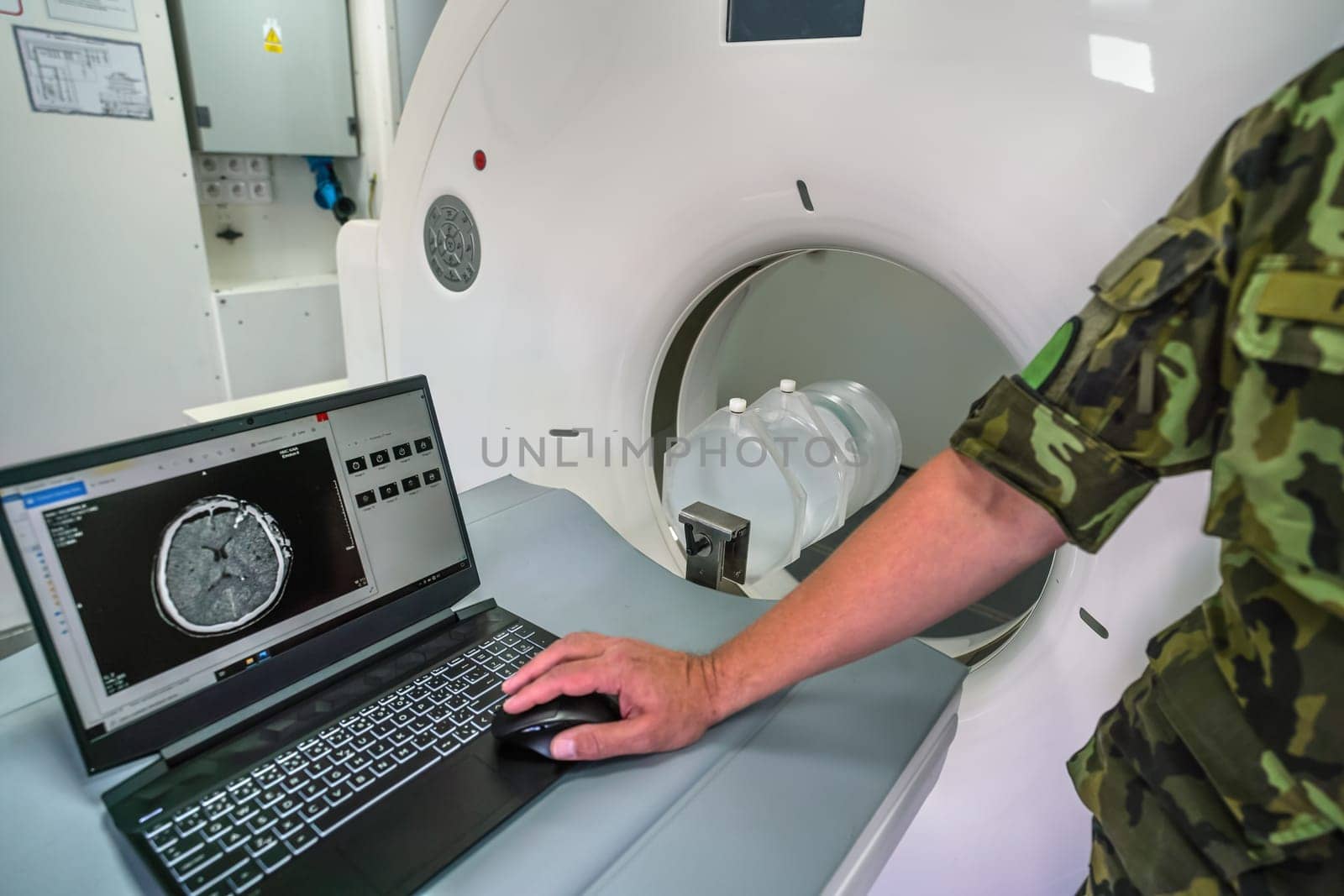 CT or computed tomography scanner inside army mobile field container ambulance, soldier showing patient with severe head trauma scan on laptop near.