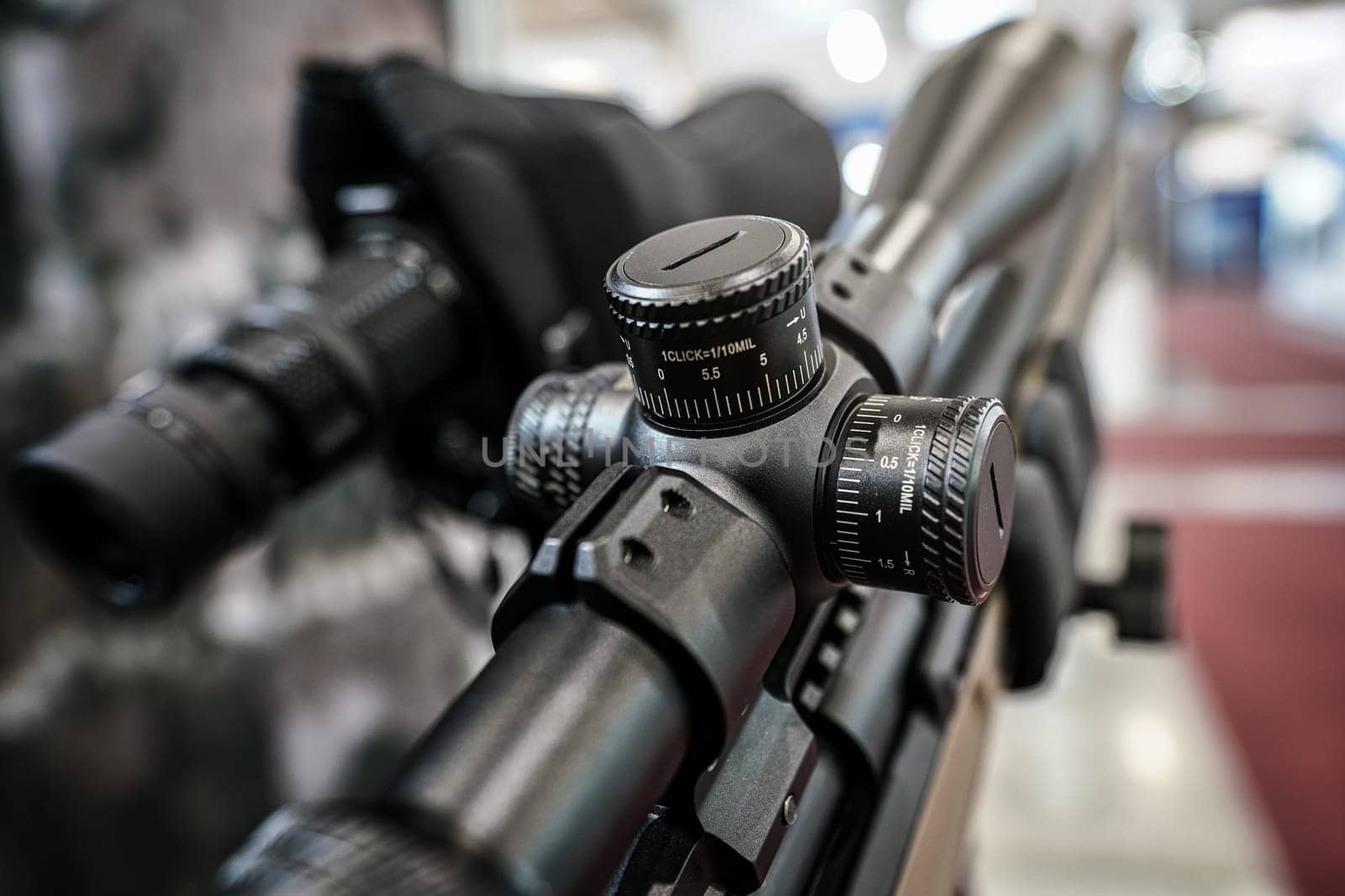 Black shooting scope optics mounted on rifle displayed at weapons exhibition fair, closeup detail to adjustment knobs.