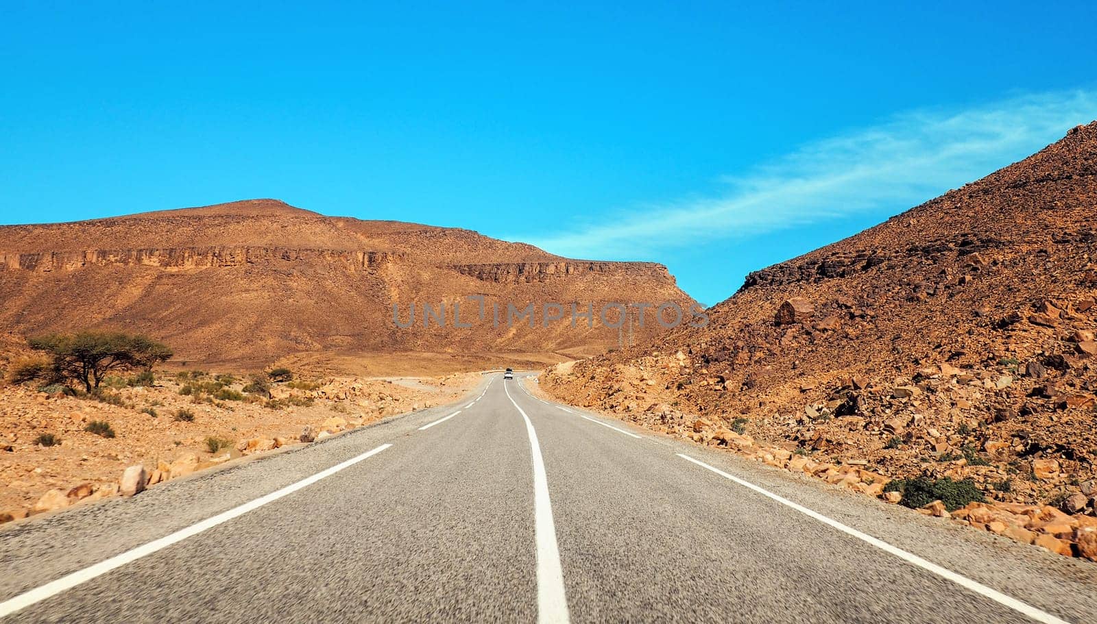 Straight asphalt road , small Atlas mountains and low bushes both sides, clear sky above - typical landscape in Southern Morocco.