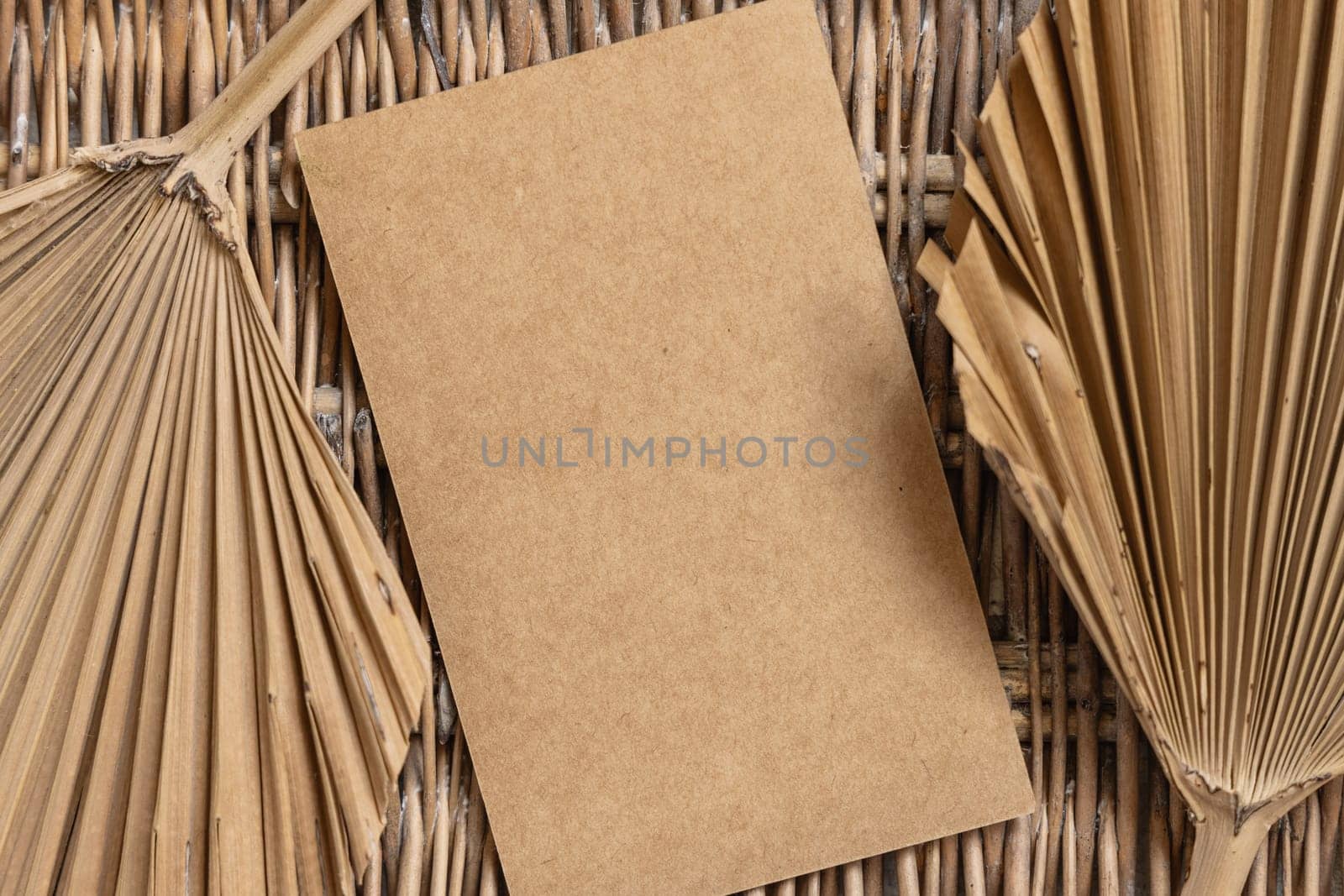 Blank card on a wattled table with dry palm leaves top view. Bohemian tropical template with vertical paper card flat lay. Greeting or Invitation card mockup, place for text