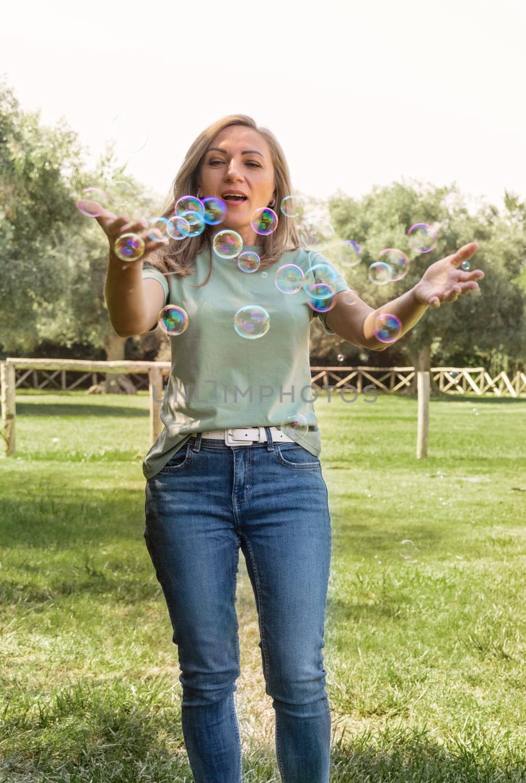 Women wearing t-shirt and jeans playing with soap bubbles in a park, three quarter shot.Middle aged caucasian lady having fun ioutdoor in a summer. Magician, outdoor activity concept.