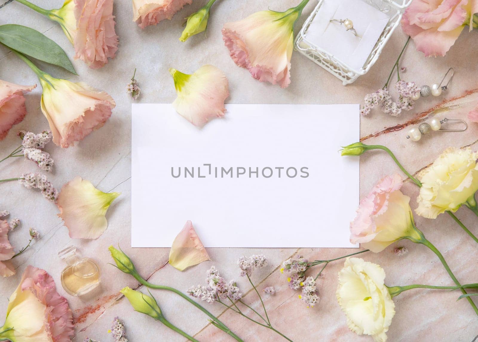 Paper card mockup between pink flowers, petals, earrings and perfume flacon top view on a marble table. Romantic scene with horizontal blank card with place for text flat lay. Valentines, Spring or Mother's day concept