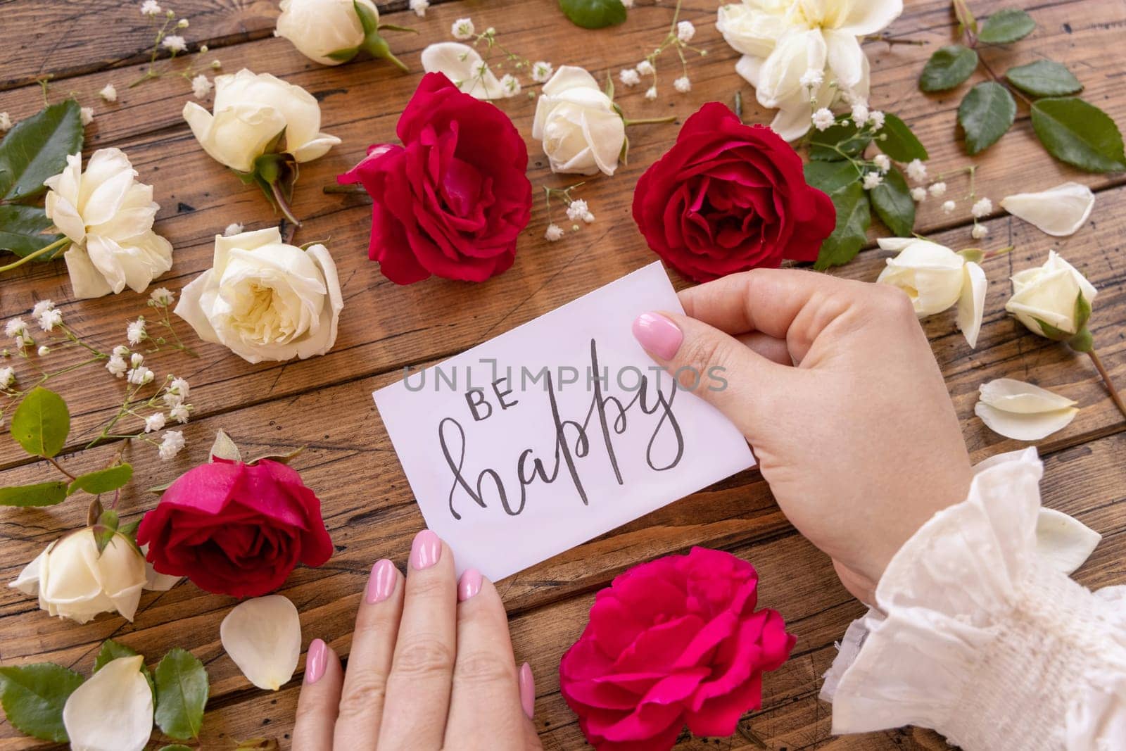 Hands with handwritten card BE HAPPY surrounded by red and cream roses close up on a wooden table. Femminine romantic declaration of love near flowers, Valentines day concept