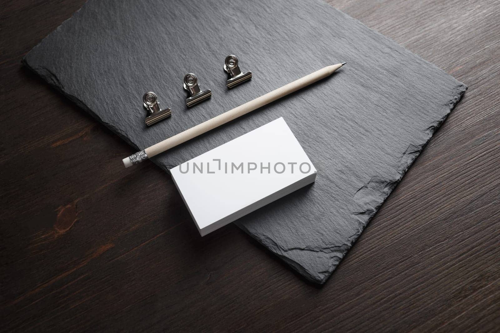 Photo of blank business cards, pencil and metal clamps on stone board background. Template for ID.