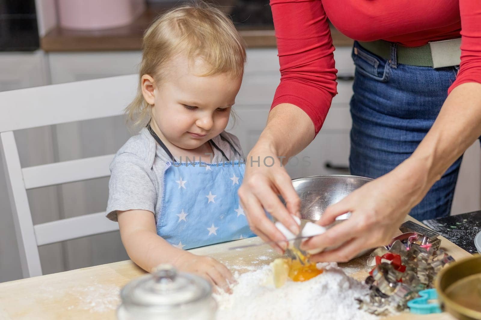 Cute boy watches mom puts eggs in flour and prepares dough in the kitchen. Horizontally.