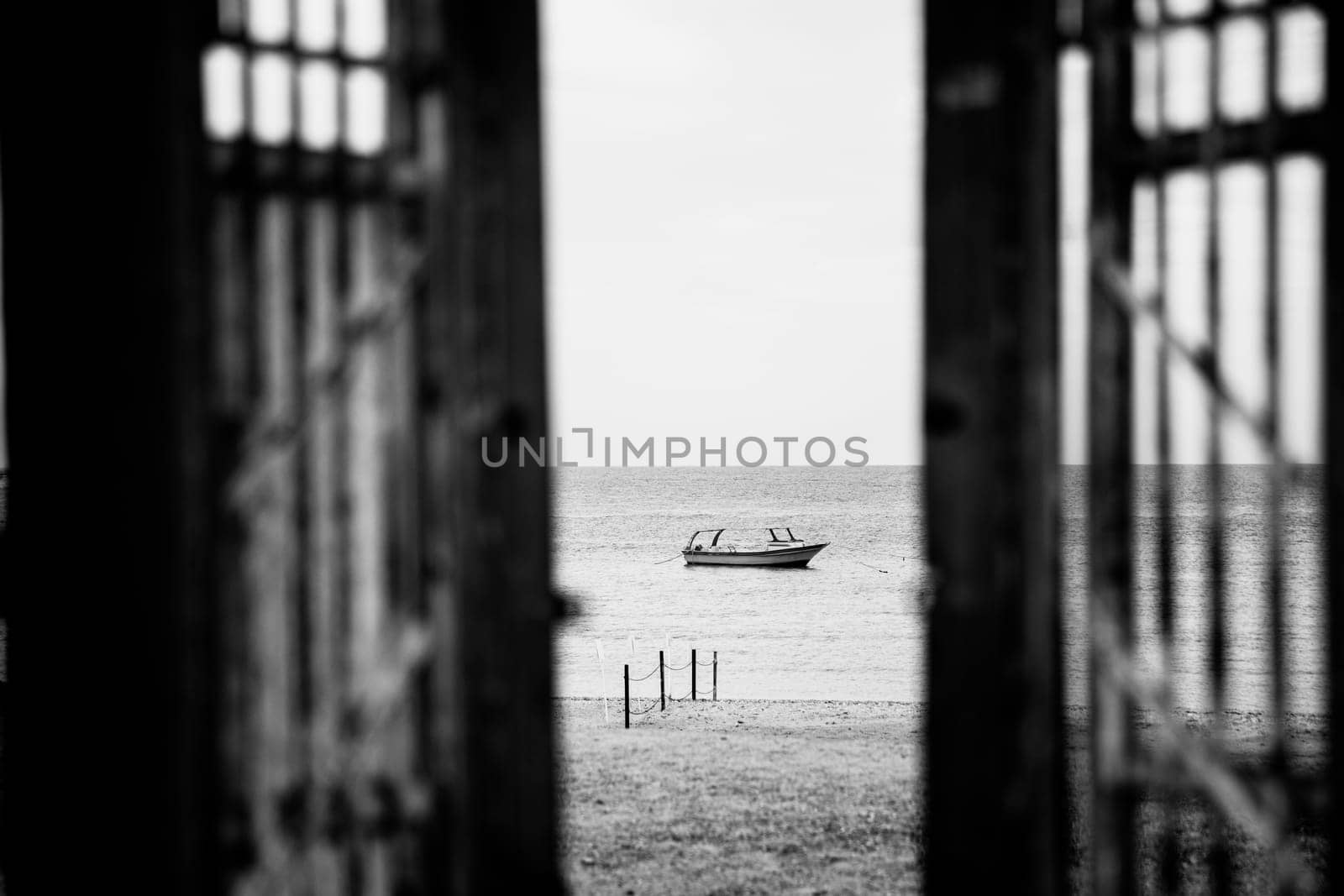 Black-white vessel or yacht photography. Opened doors to beach with alone boat or ship in silent sea or ocean water surface. Window opening to quiet and peaceful harbor or bay. No waves tranquil mood