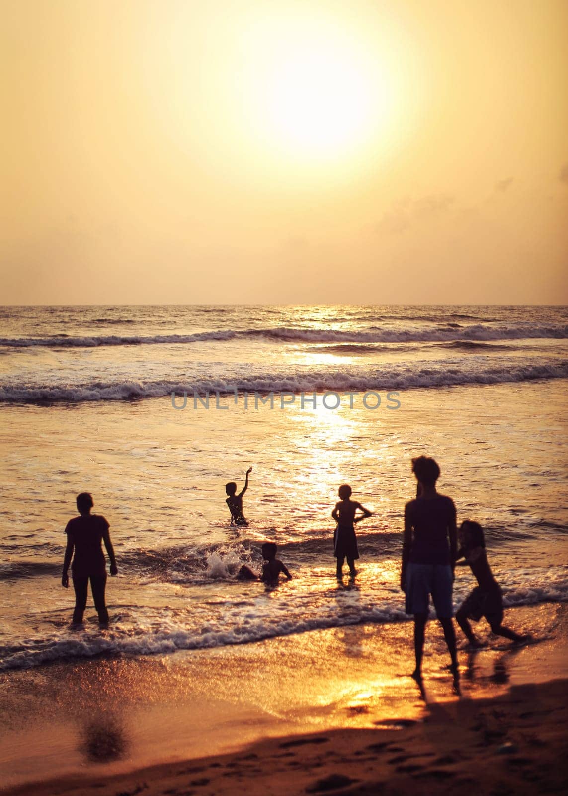 Kalutara, Sri Lanka - April 15, 2017: Silhouettes of people, mostly kids playing on the wet beach, sea and golden sunset afternoon light in back.