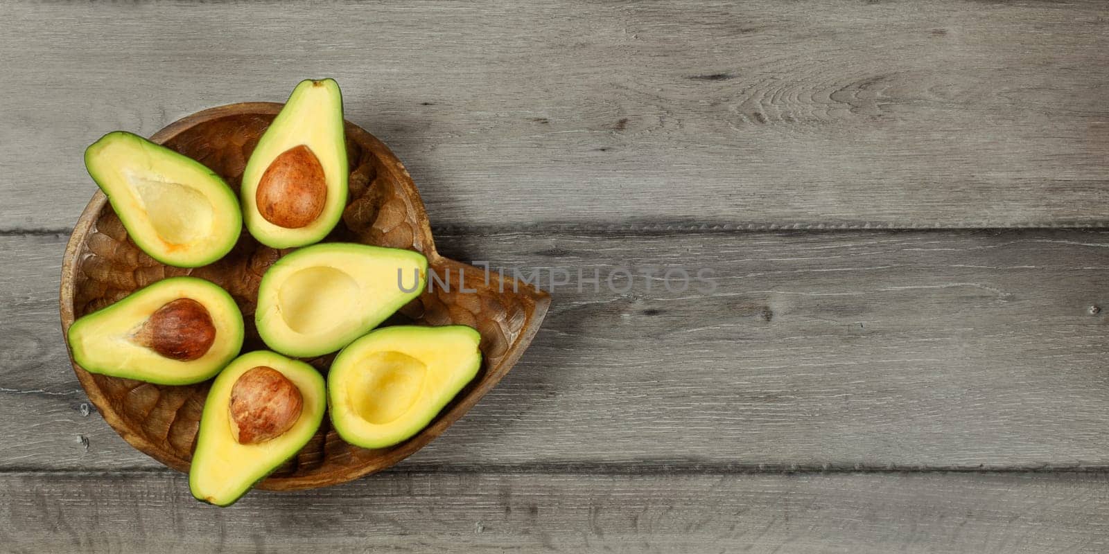 Tabletop view - avocado cut in half, arranged on wooden bowl, with dark grey wood desk under. Banner with space for text.
