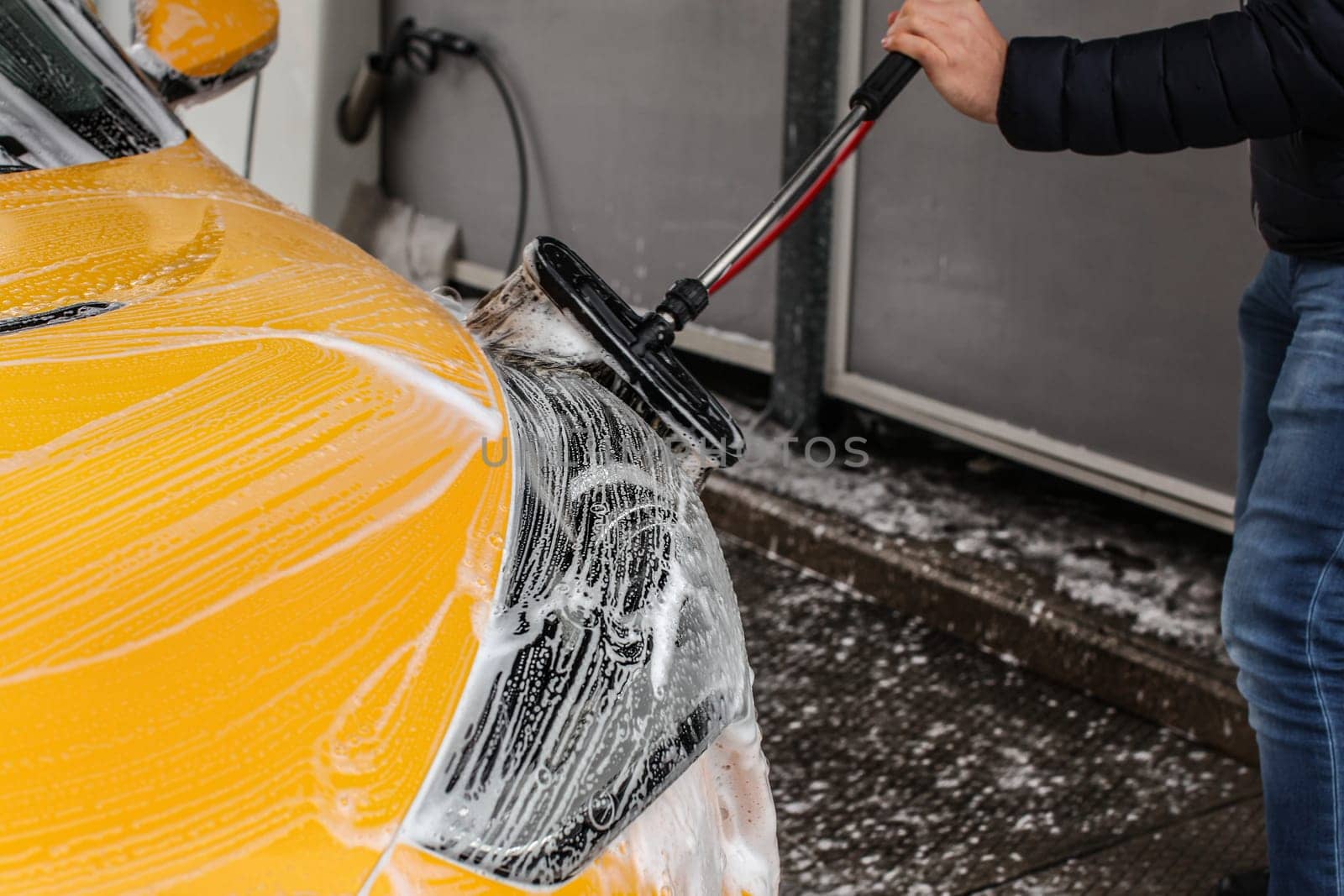 Man cleaning his car in self serve carwash, brush leaving shampoo strokes on front headlight.