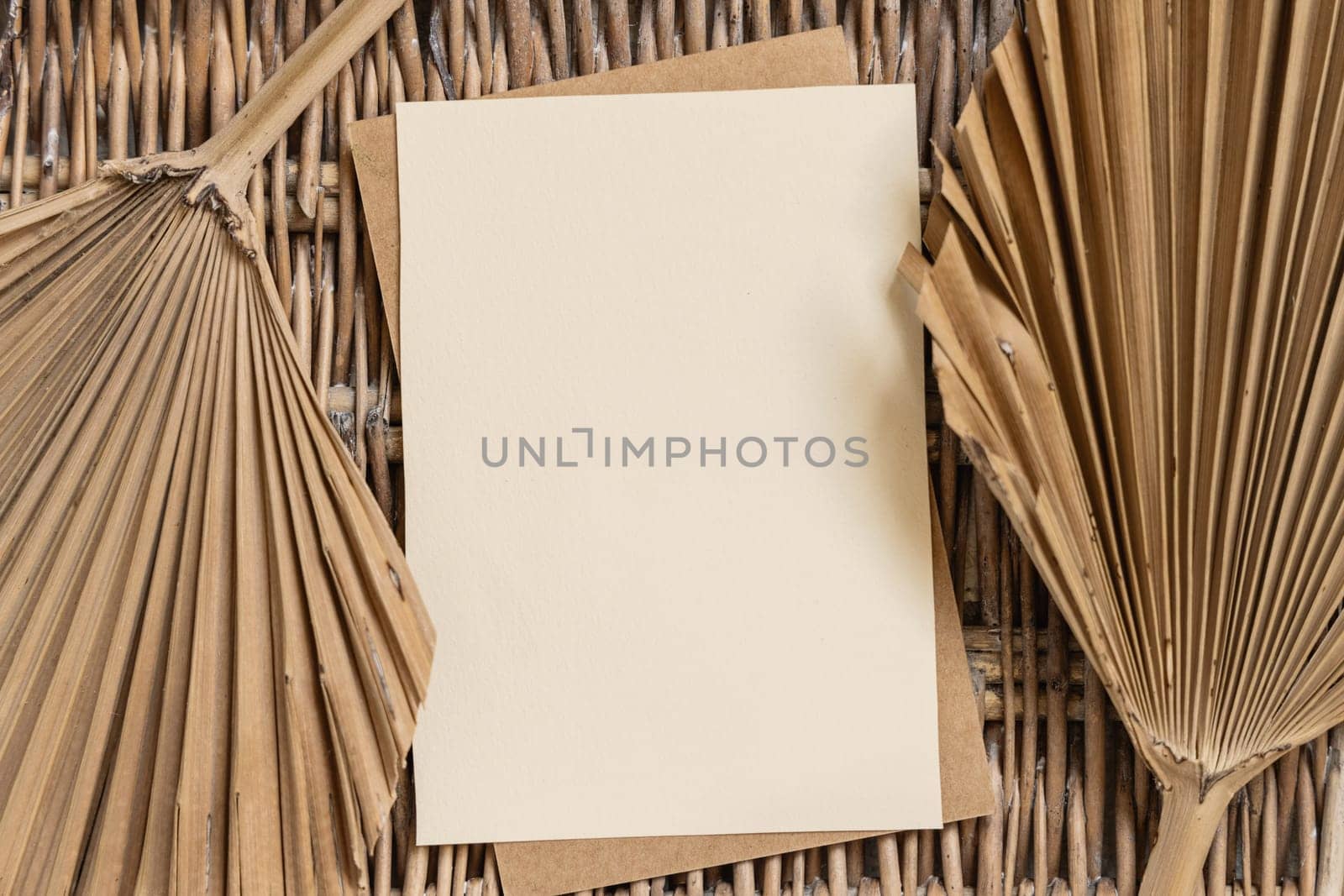Blank card on a wattled table with dry palm leaves top view. Bohemian tropical template with vertical paper card flat lay. Greeting or Invitation card mockup, place for text