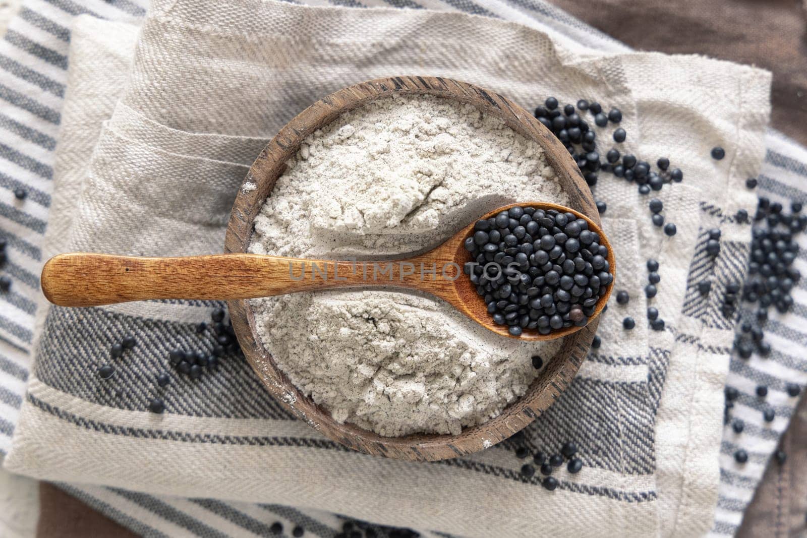Bowls with black lentils flour and spoon with beans on kitchen towels top view. Alternative flour, healthy eating and vegetarian concept. Traditional Asian and North African cuisines ingredient