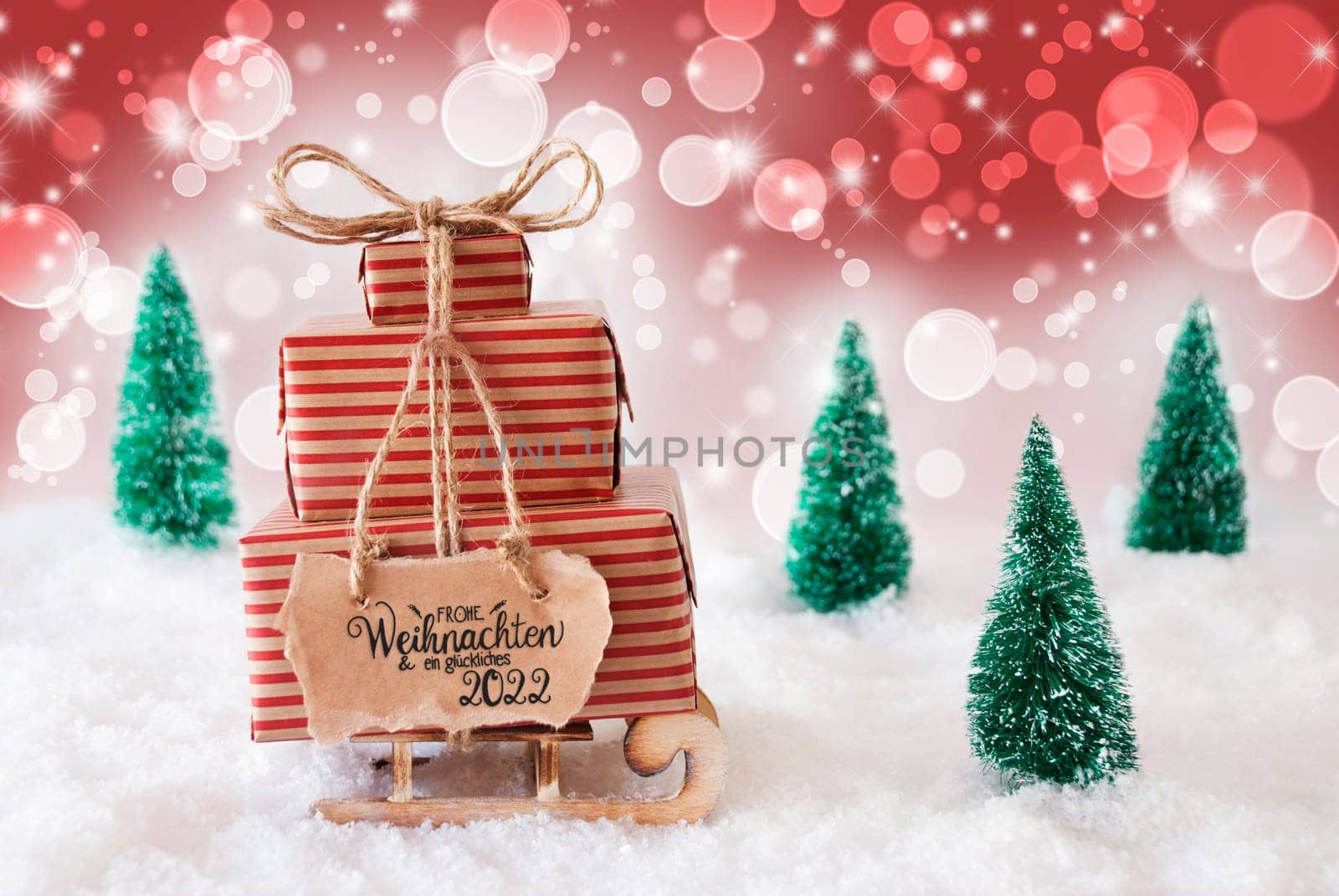 Sled With Christmas Gift And Label With German Frohe Weihnachten Und Ein Glueckliches 2022 Means Merry Christmas And A Happy 2022. Snow With Christmas Tree And Red Sparkling Background