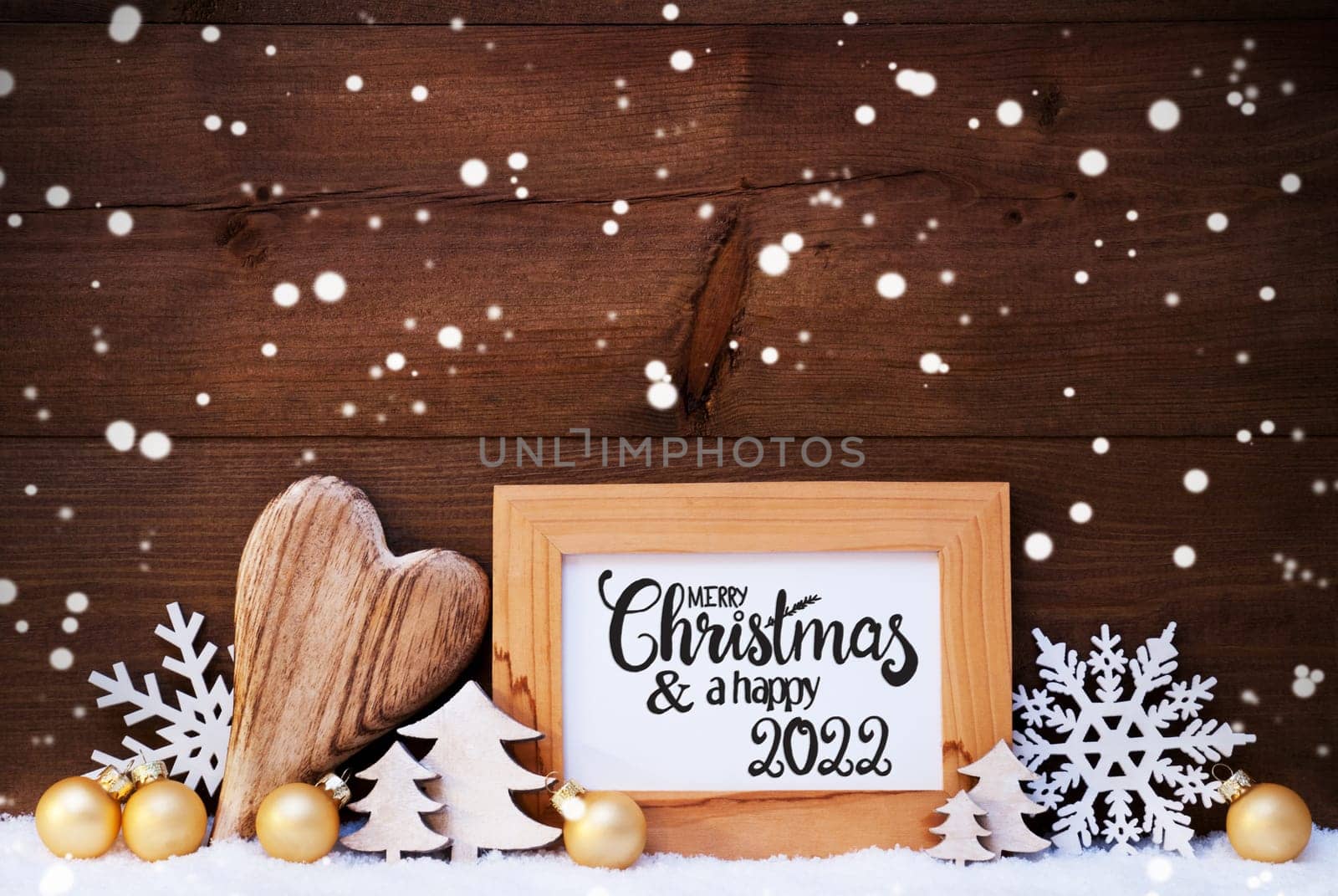 English Calligraphy Merry Christmas And Happy 2022. Golden Christmas Ornament Like Ball, Heart And Tree. Wooden Background With Snow And Snowflakes