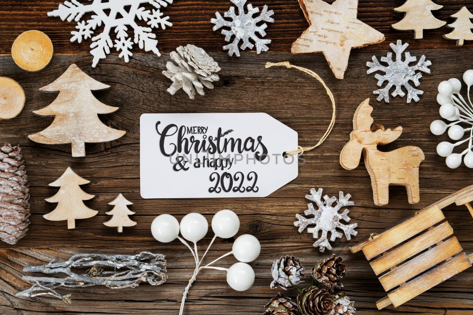 One White Label With English Text Merry Christmas And A Happy 2022. Frame Of Christmas Decoration Like Tree, Sled, Star And Fir Cone. Wooden Background With Snowflakes