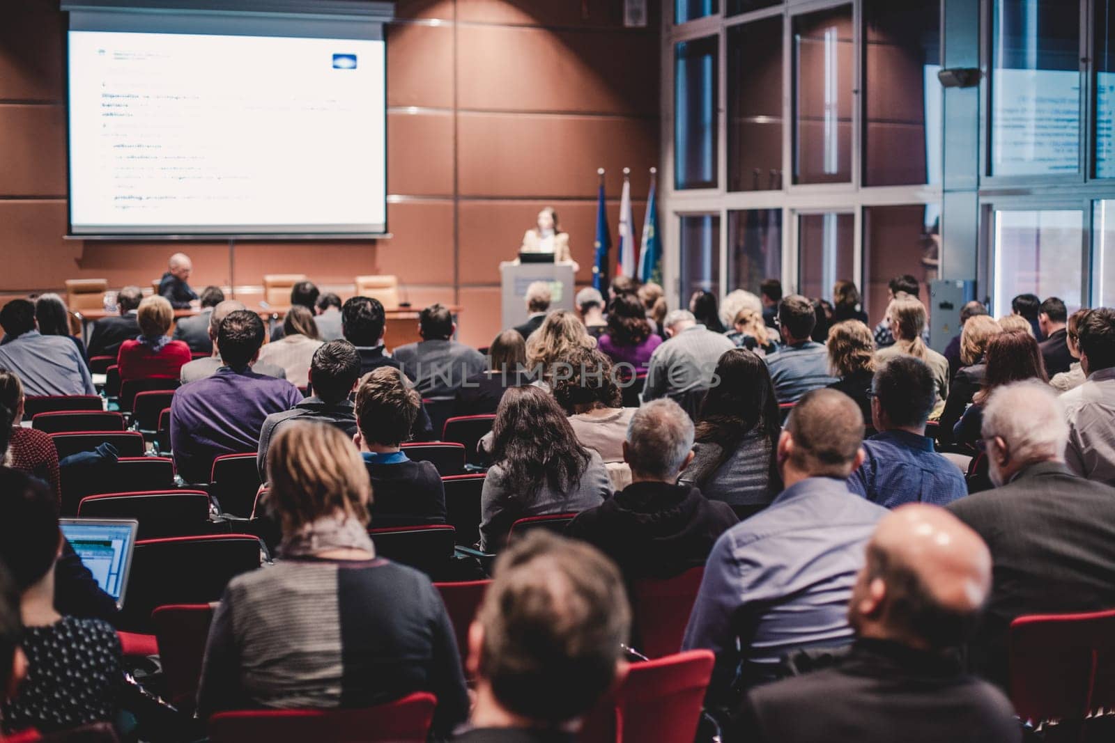 Business and entrepreneurship symposium. Speaker giving a talk at business meeting. Audience in the conference hall. Rear view of unrecognized participant in audience.