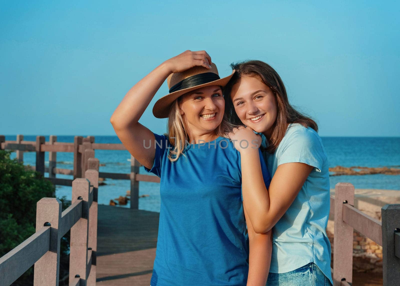 Mother wearing straw hat and teenage daughter standing on pier at sea coast, embracing and smiling, t-shirts mockup. Loving family spending time together outdoors in summer, enjoying sunset. Travel concept