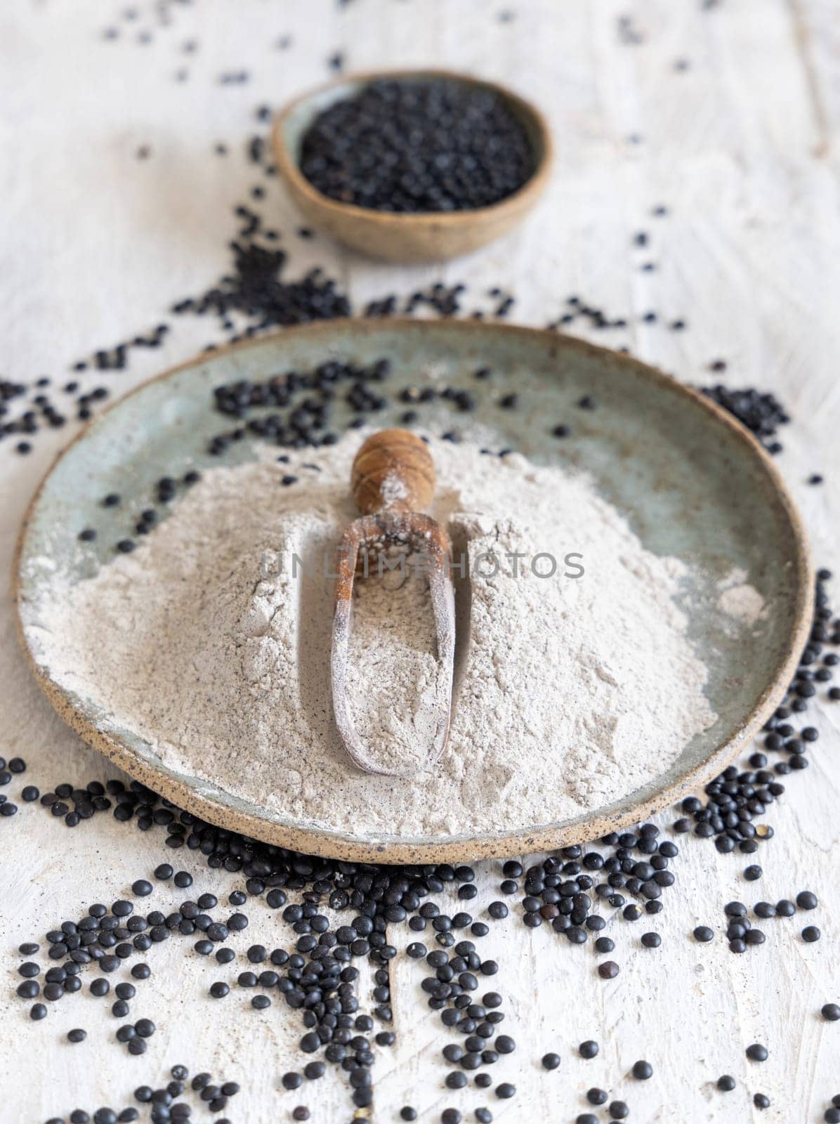 Plate with a scoop of black lentils flour and beans on white wooden table close up. Alternative flour, healthy eating and vegetarian concept. Traditional Asian and North African cuisines ingredient