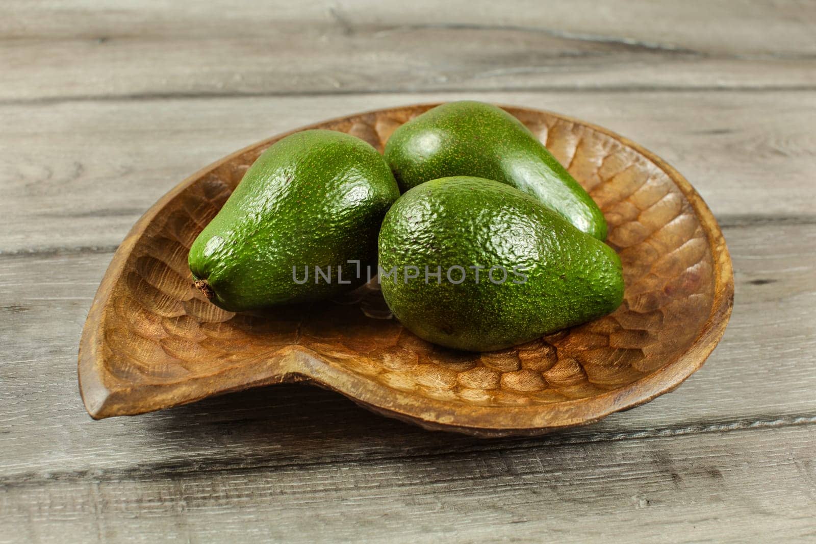 Three whole avocado pears in wooden bowl, on rustic table.