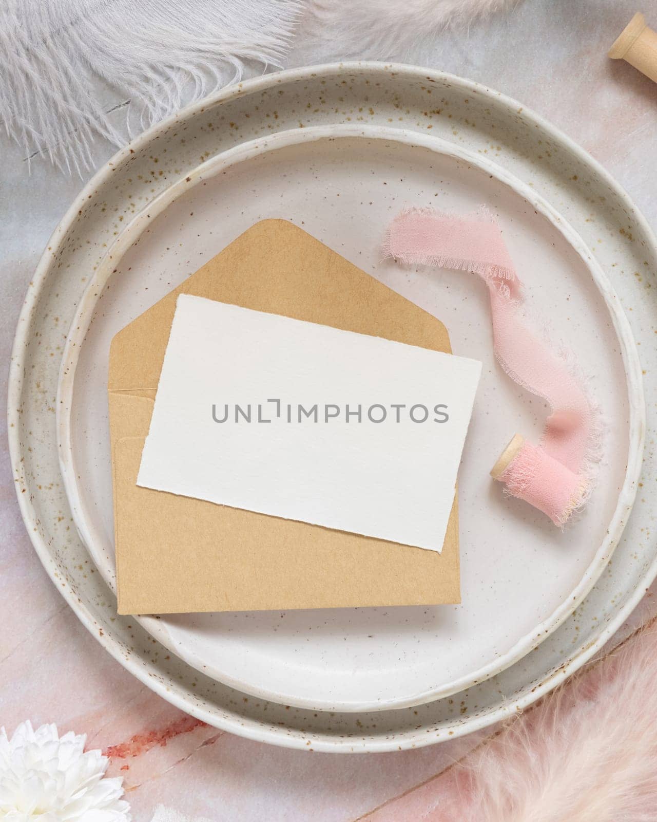 Wedding Table place with a paper card over envelope near flowers and feathers top view. Romantic template with horizontal blank card on a plate flat lay. Card mockup, copy space