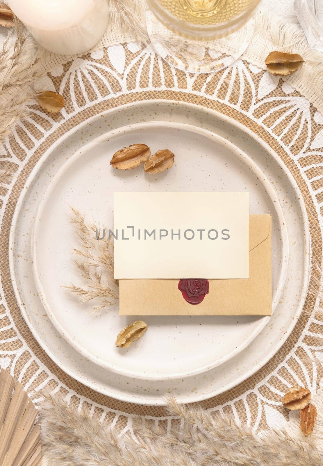 Wedding table place with horizontal blank card and sealed envelope on bohemian placemat top view. Boho wedding card mockup. Tropical beige template with dried plants and pampas grass, place fot text
