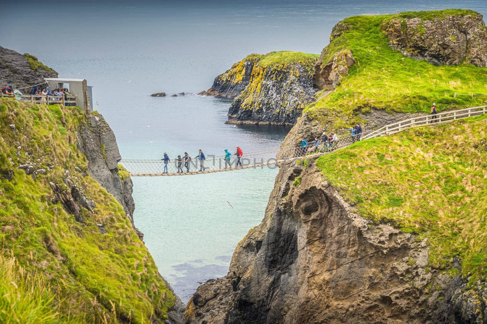 Closeup on people on the Carrick a Rede rope bridge and scenic island surrounded by turquoise Atlantic Ocean, Wild Atlantic Way, Northern Ireland