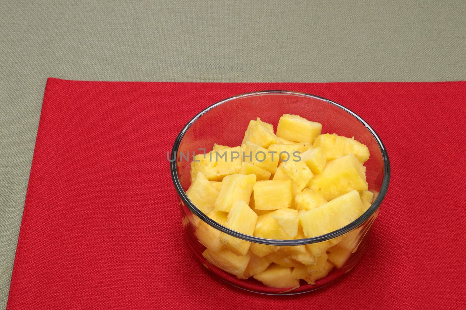 One whole sweet pineapple sliced into pieces and place in a clear glass bowl close-up on a red place mat. Sweet yellow pineapple fruit pieces in a round bowl on a red place mat on a green tablecloth.