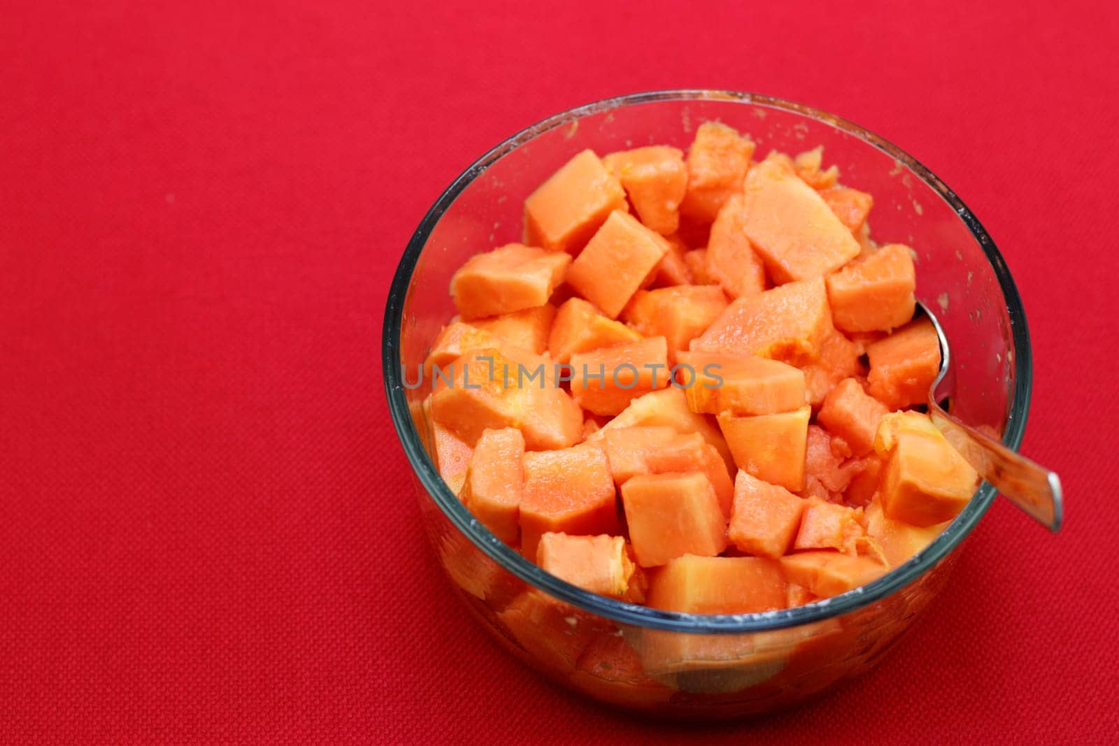 Full papaya serving of whole tropical fruit ready to eat slices prepared in a clear glass bowl with a spoon on a red background. Delicious papaya pieces ready to eat in a clear glass bowl closeup.