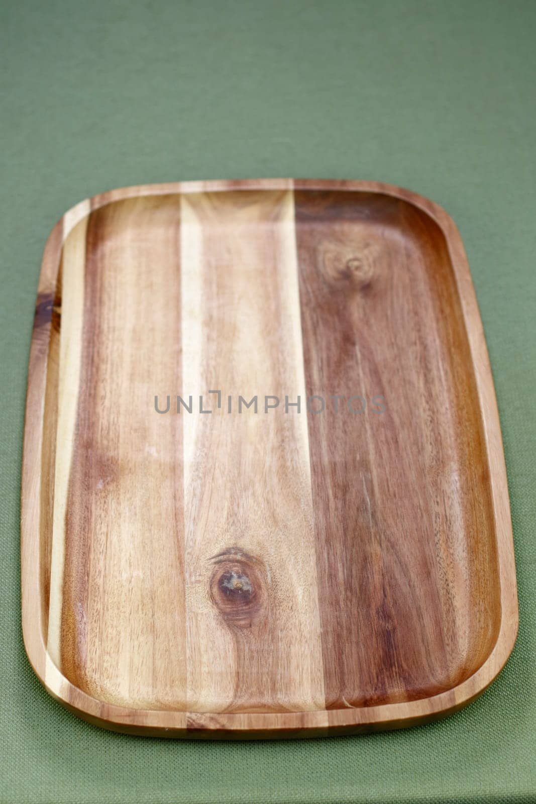 Close-up of a hardwood beige and brown rectangle serving tray with a rail or lip raised edge. Vertical acacia wooden rectangle tray that is used to serve food resting on a green table cloth close-up.