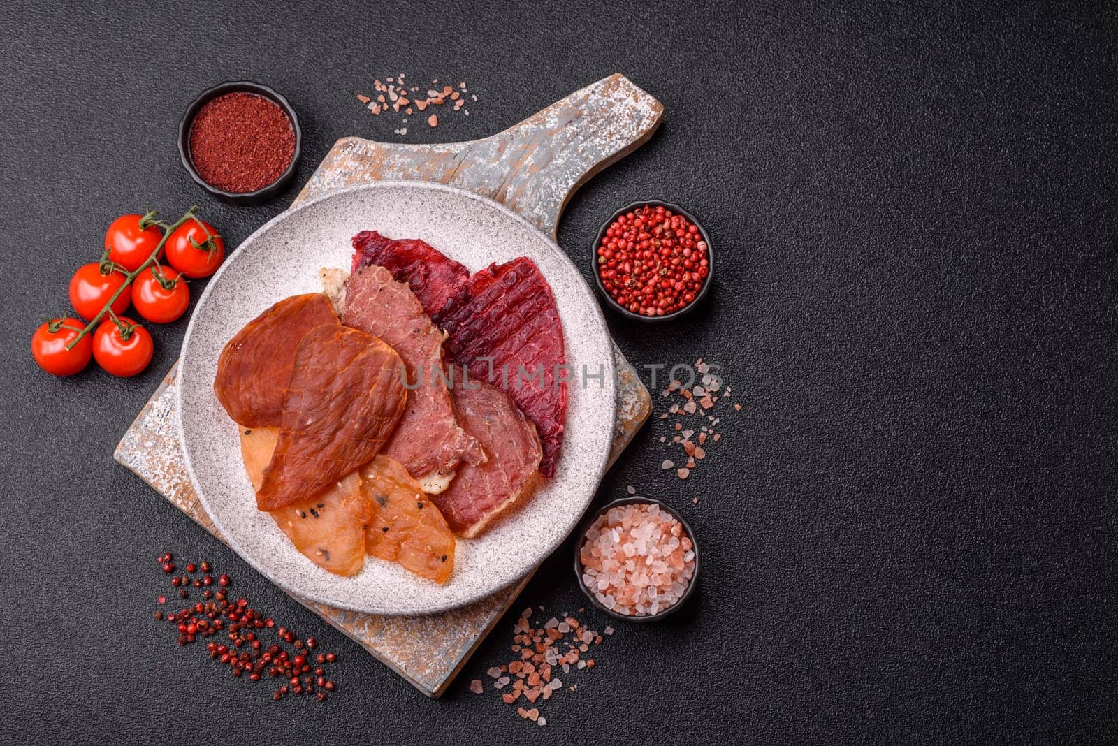 Jerky, dried meat in the form of slices with salt, spices and herbs on a dark concrete background
