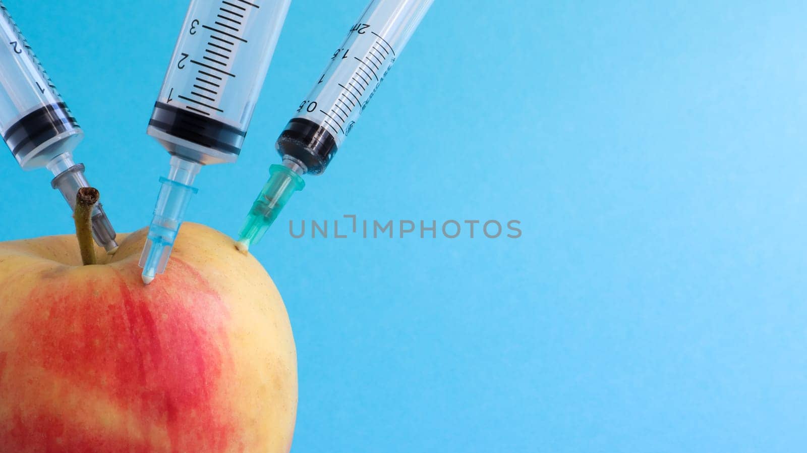 Three different medical syringes sticking deep into an apple on a blue background. The fruit is strewn with syringes. A ripe red apple pierced from all sides by syringes. Healthcare or cosmetology
