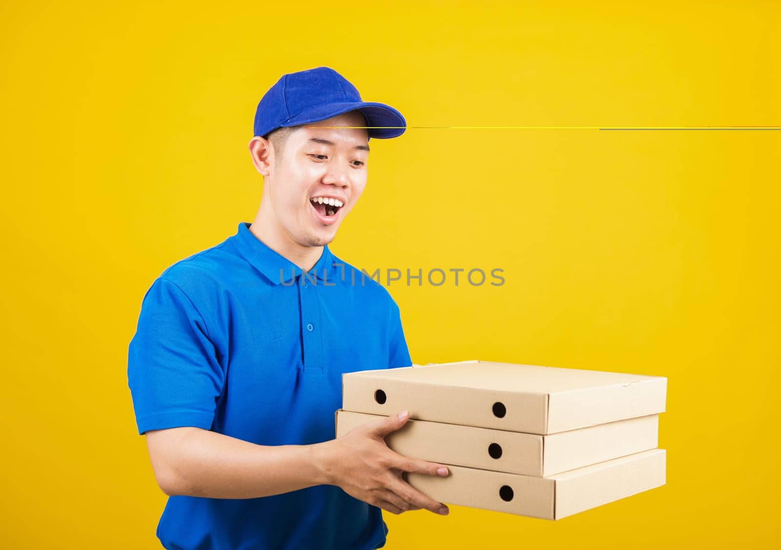 Portrait excited delivery service man standing he smile wearing blue t-shirt and cap uniform hold give food order pizza cardboard boxes looking to camera, studio shot isolated on yellow background