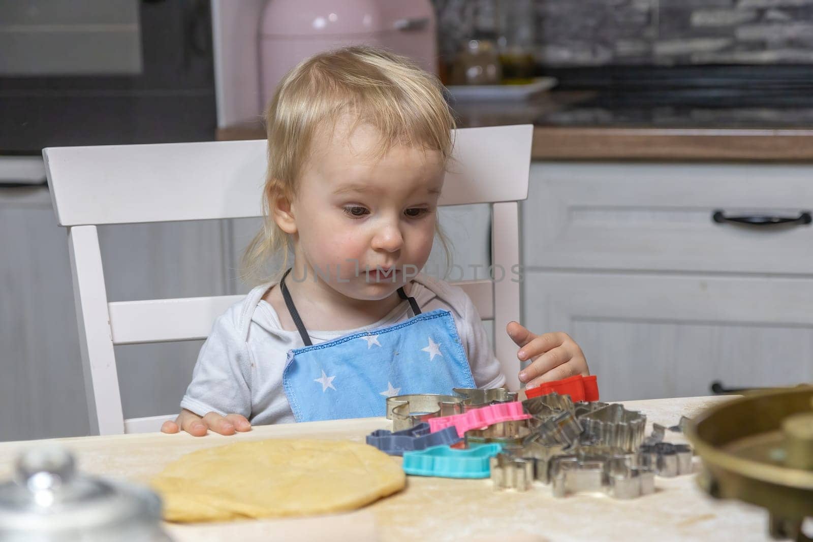 Funny blond little child boy looks at the dough cutters in surprise in domestic kitchen. Horizontally.