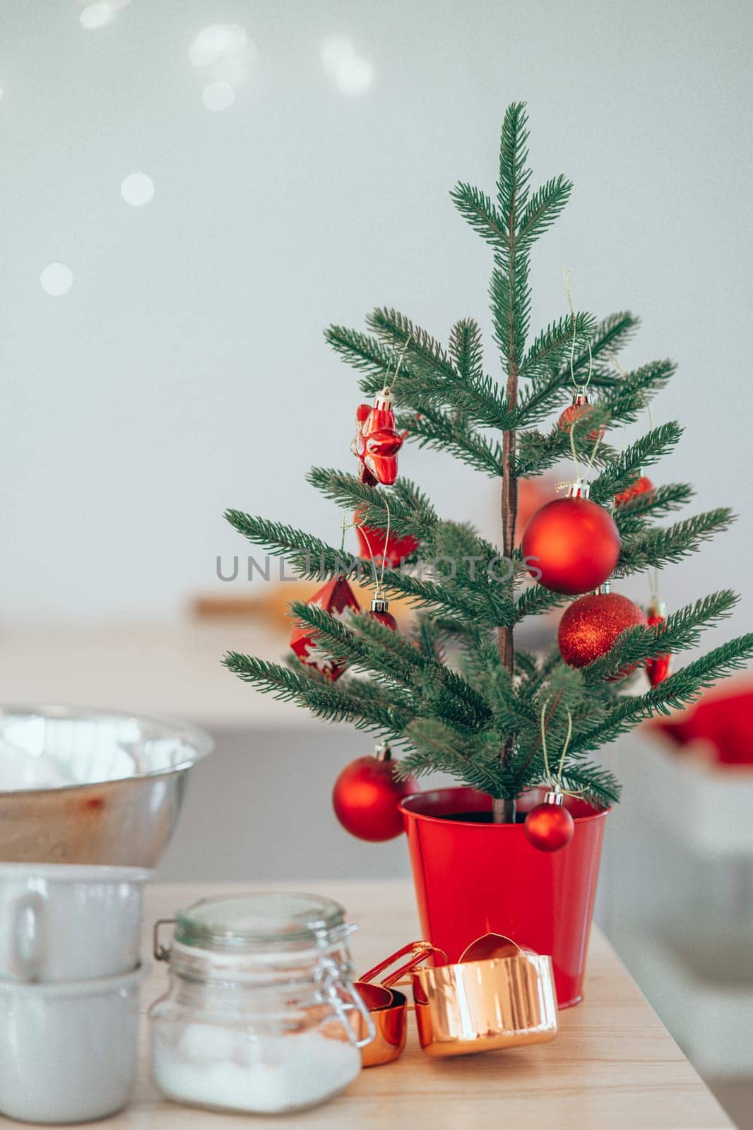 Small decorated Christmas Tree with Christmas toys in red pot placed on kitchen table near cups and sugar. Christmas decoration in the kitchen at home. Concept of celebration of Christmas Holidays.