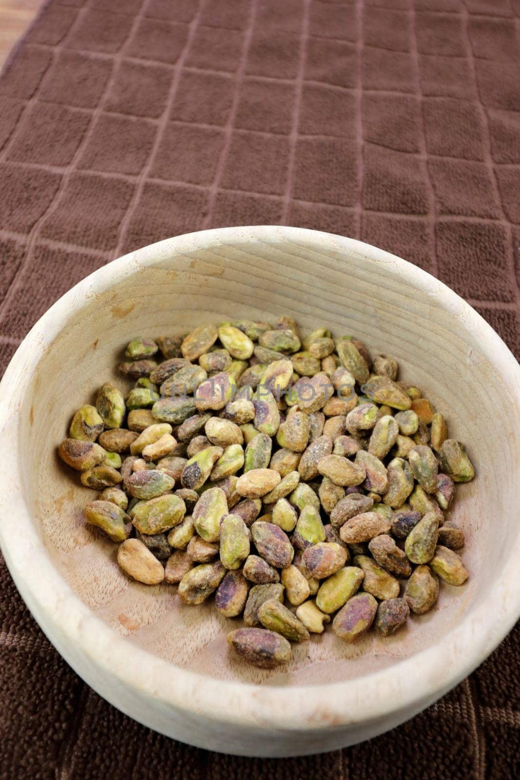 Round wooden bowl of green roasted pistachio nuts without shells closeup on a brown towel on a table. Close-up of shelled roasted pistachio nuts in a wooden bowl resting on a brown towel on a table