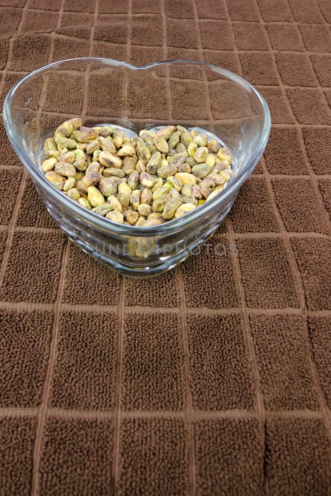 Roasted shell-less pistachios with sea salt in a heart shaped clear glass dish on a brown towel with rectangle pattern. On a brown towel is a heart shaped clear glass dish with salted pistachios food