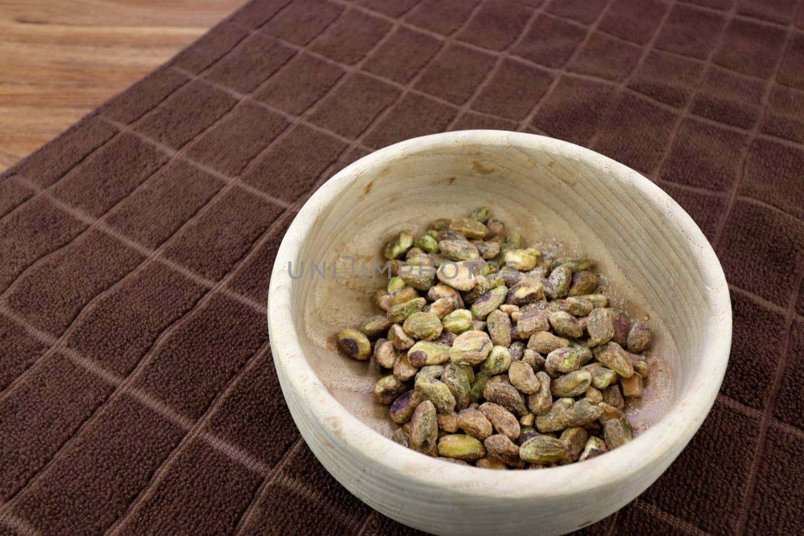 Shelled pistachio nut with sea salt in a round wood bowl on a brown towel close-up. Delicious pistachios and sea salt without shells closeup in a round wooden bowl on a brown towel resting on a table.
