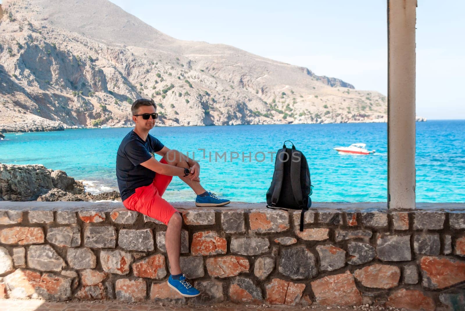 A man in pink shots, a T-shirt and sunglasses sits on a parapet by the sea.