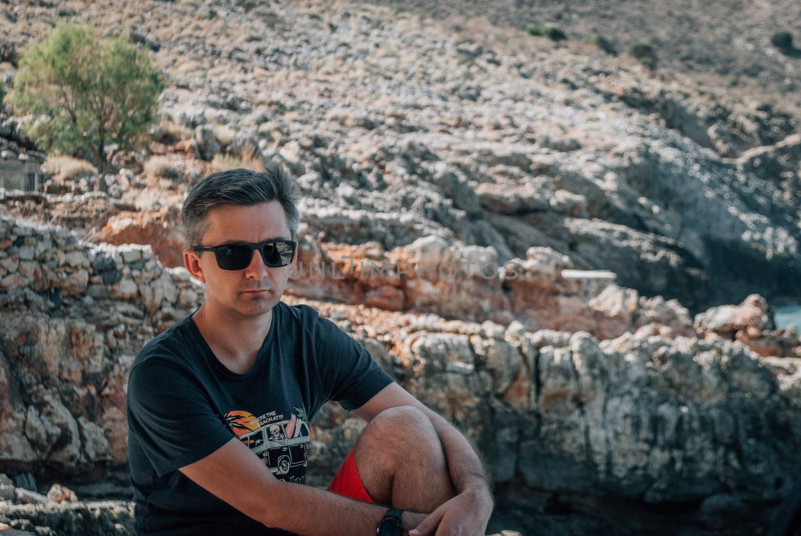 A man sits and hugs his knee against the backdrop of the rocky mountains.