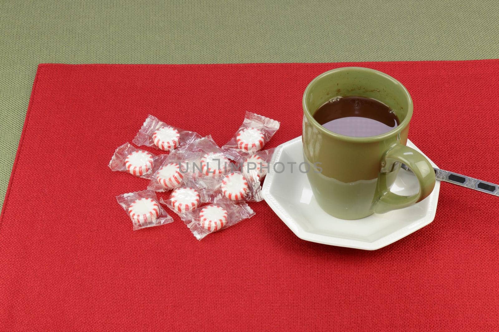 Hot cocoa beverage of a chocolate holiday mix served in a green coffee cup near peppermint candy on a red placemat on a green tablecloth. Hot chocolate cocoa mix beverage with peppermint candy rounds..